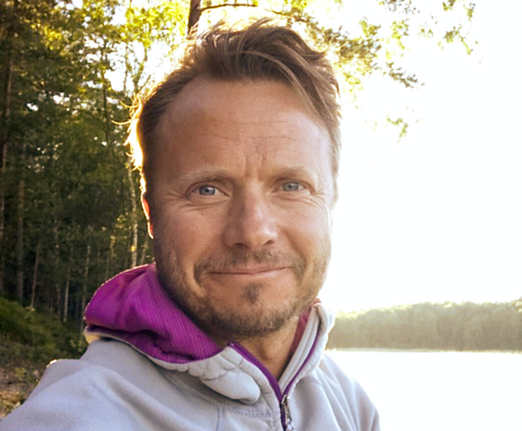 Anders står i skogen vid en sjö och ler vänligt iförd en vit fleece-jacka.