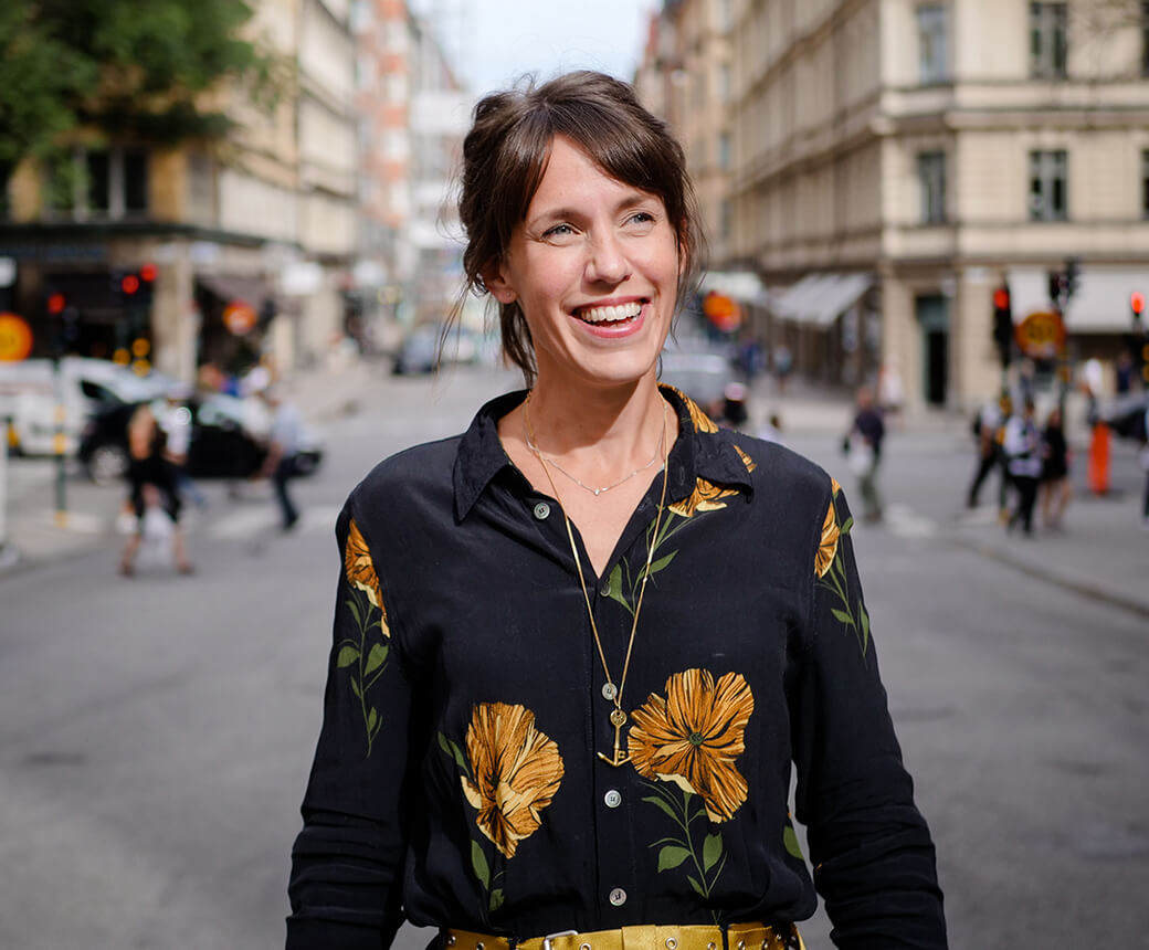 Märta är utomhus på en livfull gata. Hon ler och har en svart blus med stora gula blommor.
