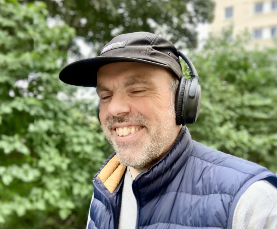 Martin har svart keps, blå väst och hörlurar. Han ler åt något som sägs i hörlurarna. I bakgrunden syns buskar och träd.