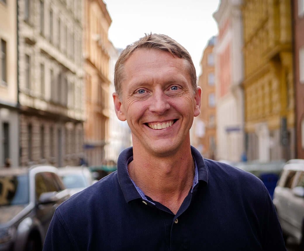 Per ler glatt och bär en mörkblå tröja. Han står mitt i en stadsgata.